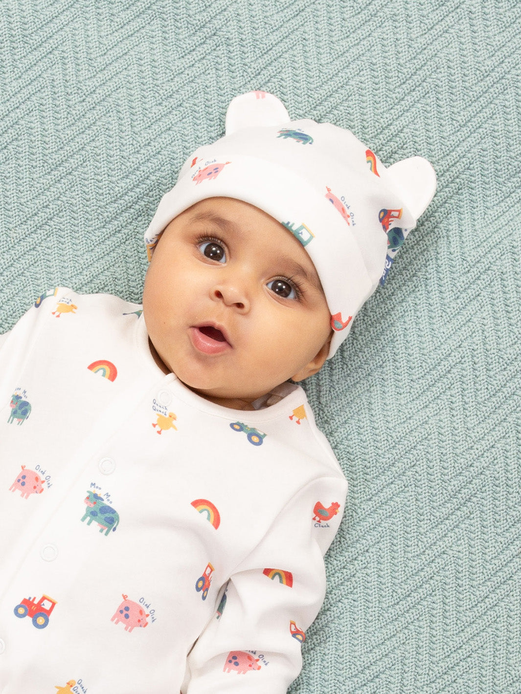 Baby wearing a white hat and matching outfit with colorful patterns on a textured green background