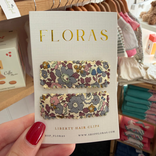 A hand with red nail polish holds a display card featuring two floral hair clips. The card is branded FLORAS and reads 'LIBERTY HAIR CLIPS.

Hair Clips
Liberty Print
Gift inspiration 
Floral
Girls Hair Accessories