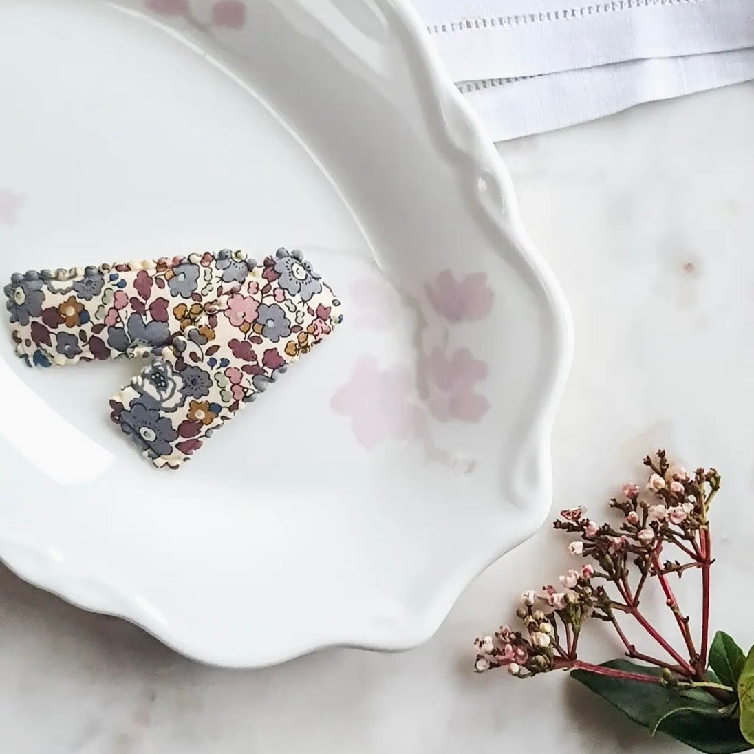 Two fabric hair clips with a multicolor floral pattern resting on a white ceramic dish, styled on a marble surface with dried flowers.

Hair Clips
Liberty Print
Gift inspiration 
Floral
Girls Hair Accessories