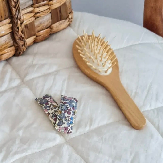A wooden hairbrush with cream-coloured bristles and a floral fabric-covered handle lies on a white quilted blanket, alongside two matching fabric hair ties with a blue and pink floral pattern.

Hair Clips
Liberty Print
Gift inspiration 
Floral
Girls Hair Accessories