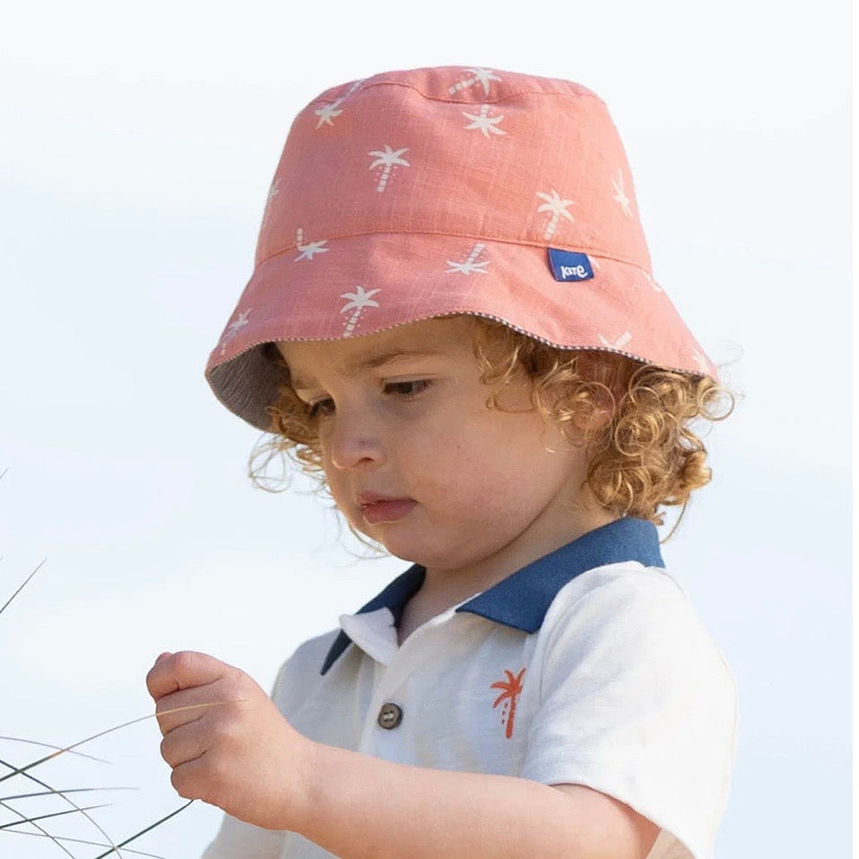 A toddler wearing an orange bucket hat with a white palm tree pattern, paired with a white polo shirt featuring a navy collar and a small red palm tree graphic. The child is looking down while outdoors.

Sun Hat
Children's clothing 
Children's sun hat 
GOTS Certified fabrics 
Reversible 