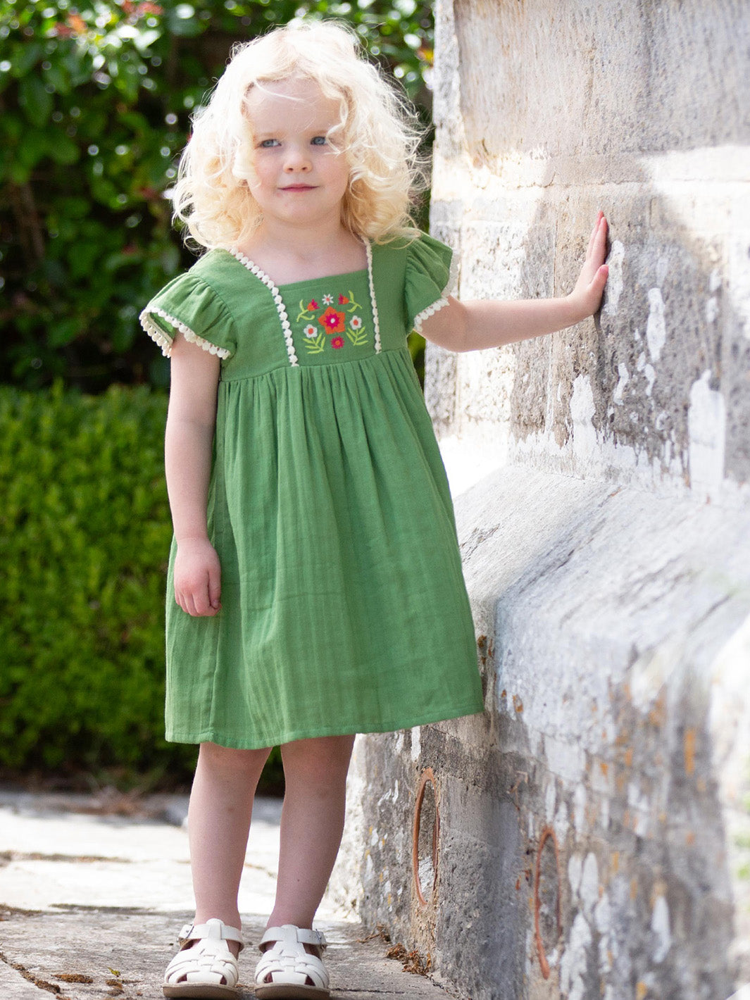 Young girl in a green dress with floral embroidery standing against a stone wall

Children's clothing 
Organic cotton 
Fairy 
Dress
Summer Clothing 
Summer outfit