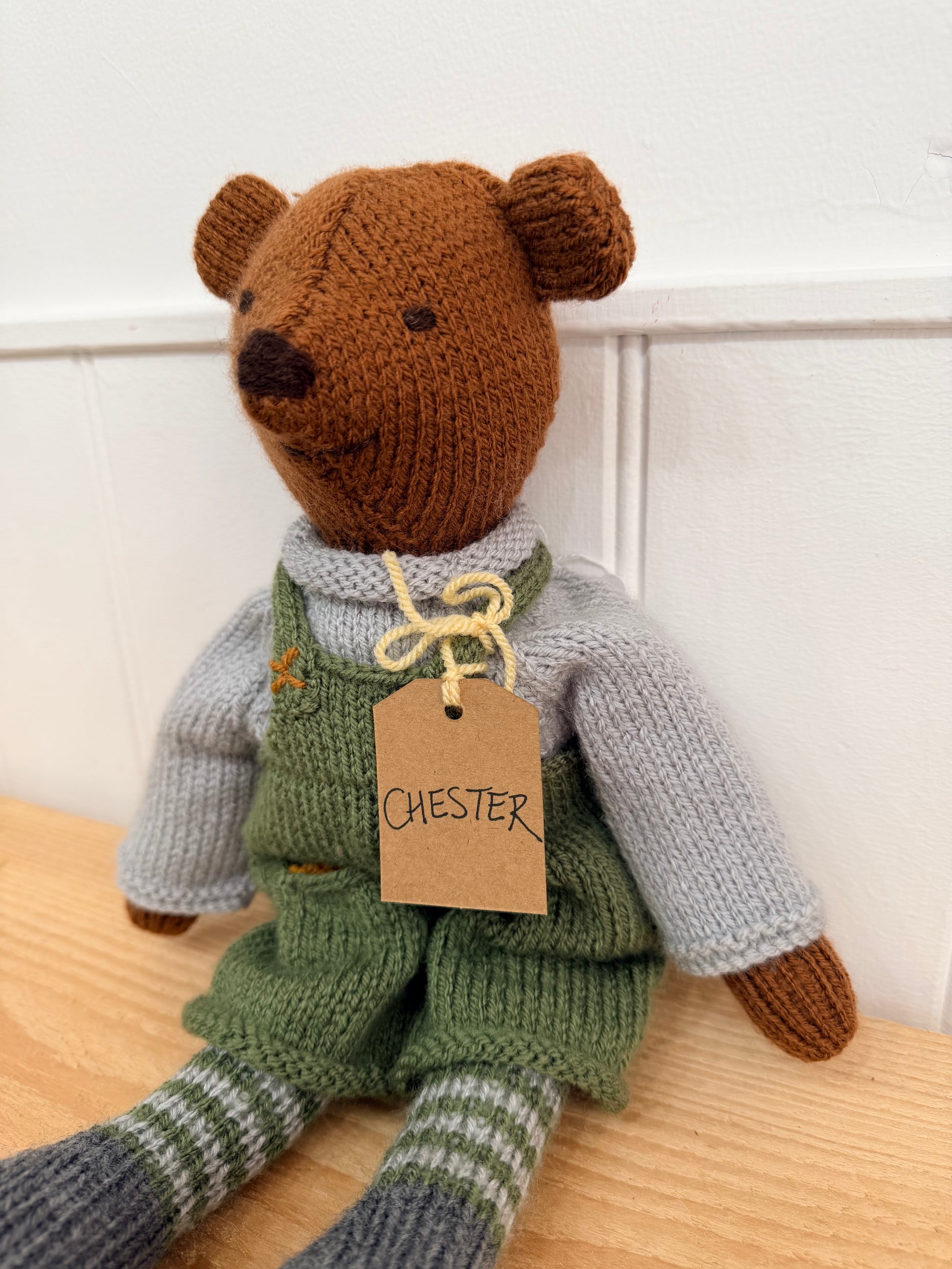 A handmade knitted teddy bear named Chester, wearing green overalls, a light grey sweater, and striped socks, sitting on a wooden surface against a white panelled wall.

Hand knitted bear 
Perfect for new baby presents
Locally made
Hand made
Unique presents 