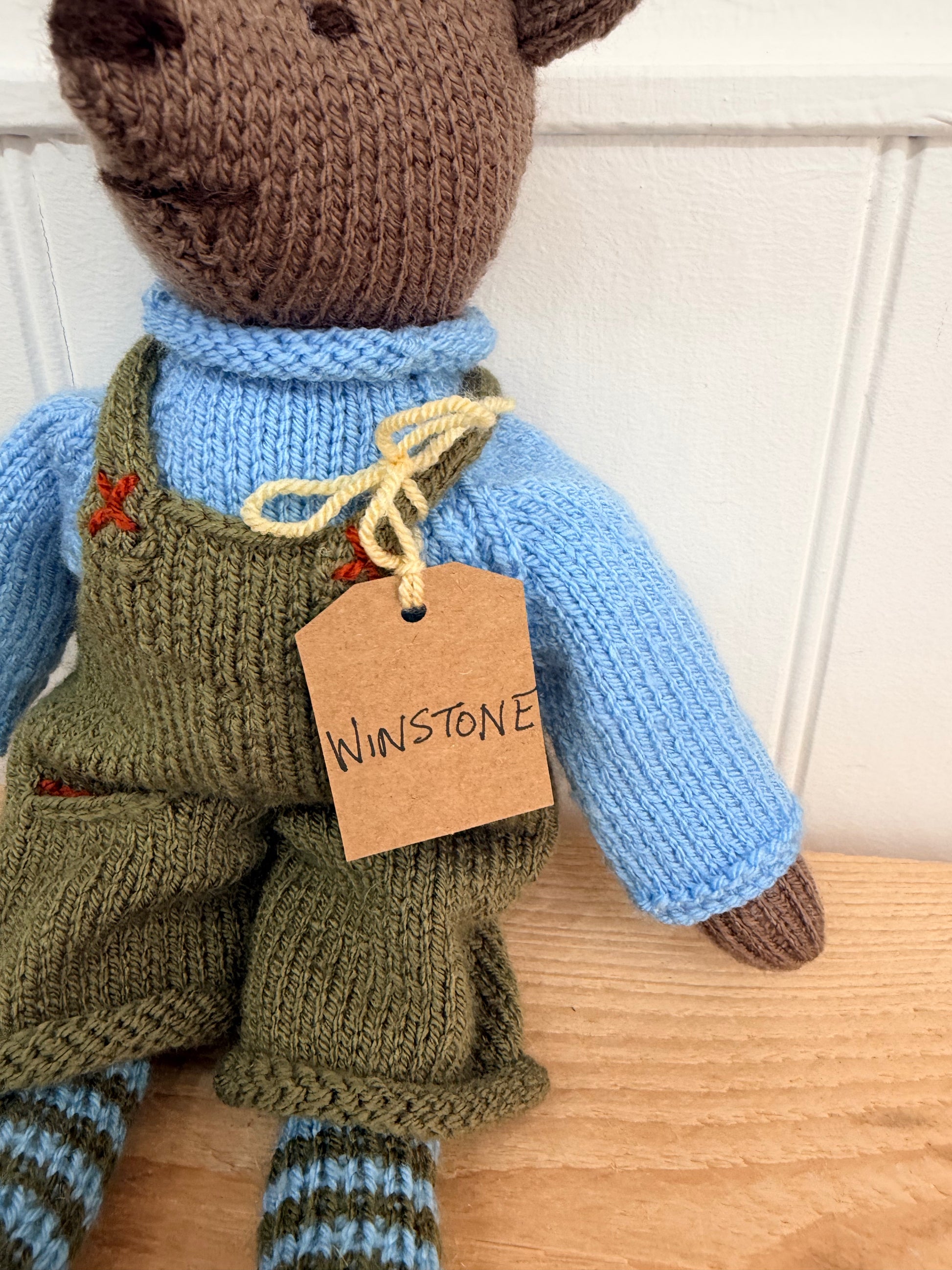 A close-up of a hand-knit teddy bear doll wearing a light blue turtleneck sweater and olive green overalls, with a brown paper tag reading 'Winstone' tied to its neck, sitting on a wooden surface against a white panelled wall.

Hand knitted bear 
Perfect for new baby presents
Locally made
Hand made
Unique presents 