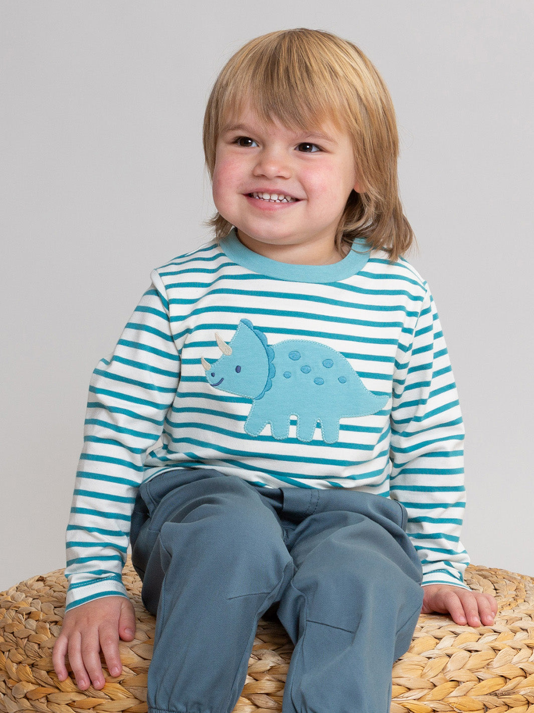 A smiling young child with blonde hair sits on a woven straw stool, wearing a long-sleeve shirt with teal and white horizontal stripes and a blue dinosaur appliqué on the front, paired with slate blue pants.

Children's clothing
T-Shirt
Organic cotton 
Dino
Boys clothing
Kite clothing