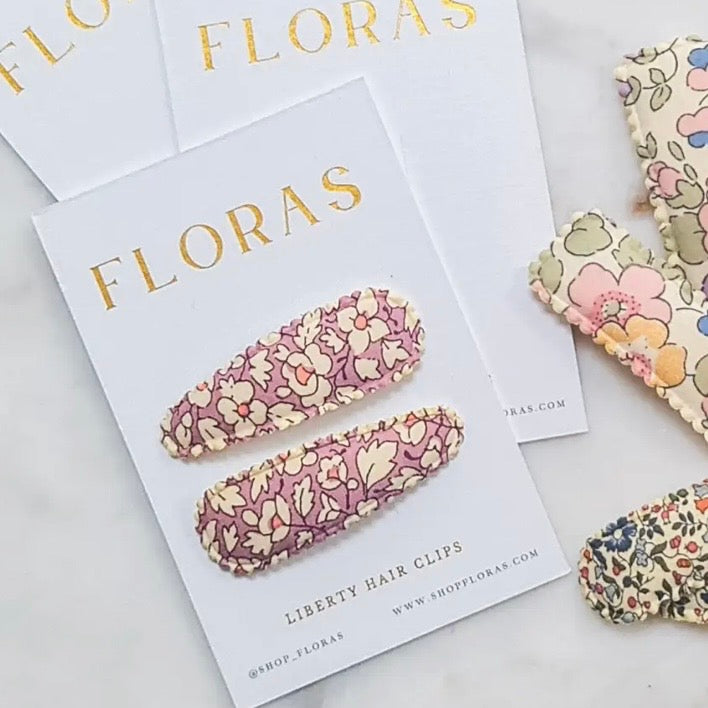 Two floral Liberty Hair Clips displayed on a white card labeled FLORAS, with additional matching floral fabric pieces arranged around them on a light surface.

Hair Clips
Liberty Print
Gift inspiration 
Floral
Girls Hair Accessories