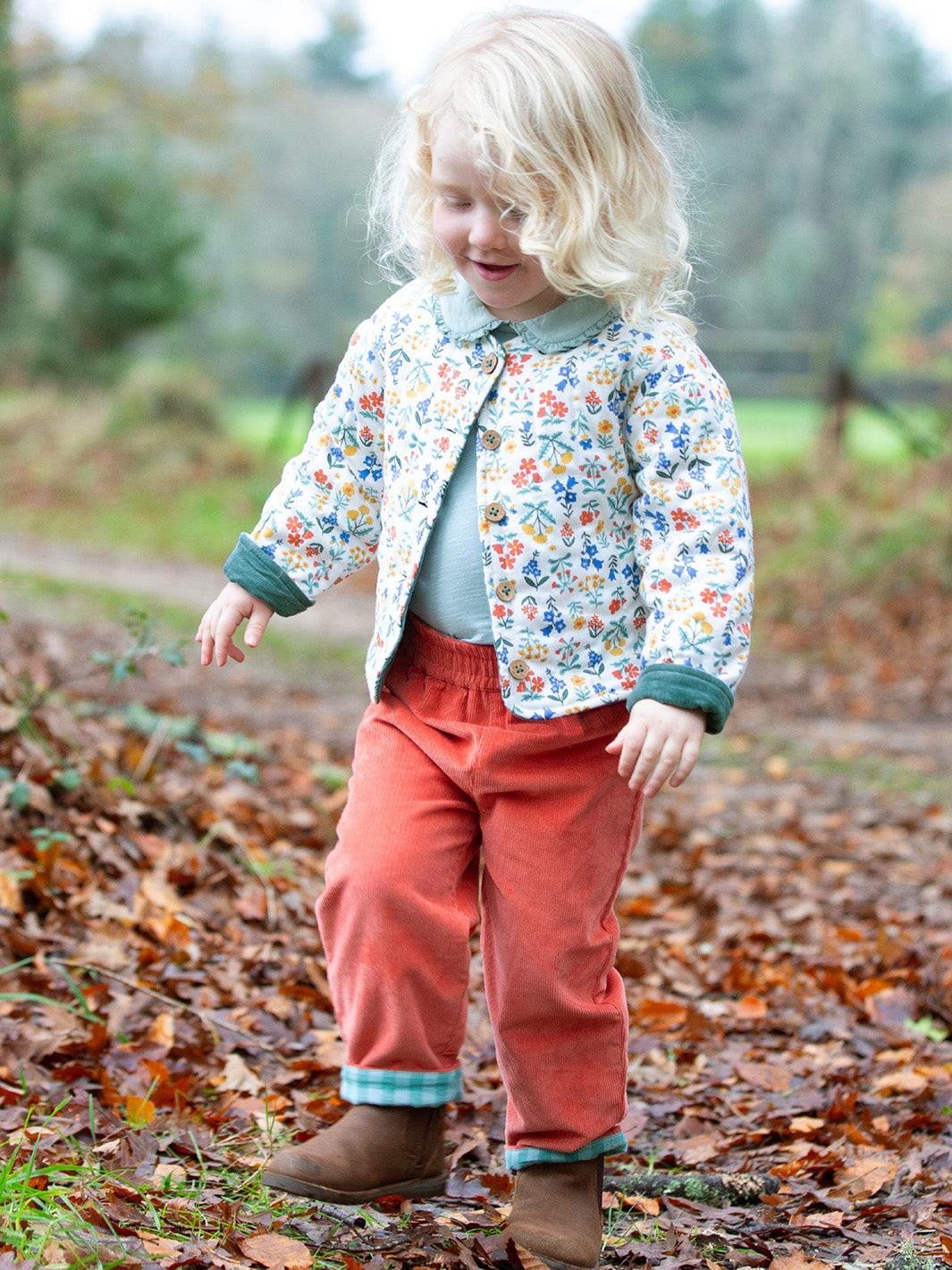 Child wearing a floral jacket and orange cords standing on a path with fallen leaves.