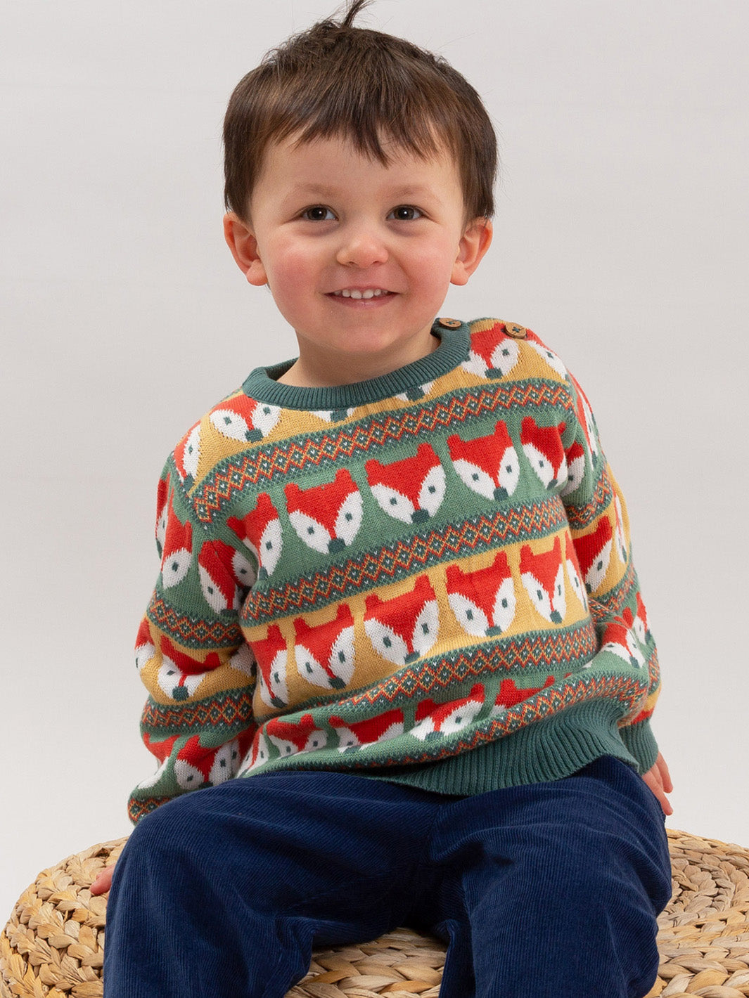 A smiling young boy wearing a colourful knit sweater with a repeating fox pattern, sitting on a woven straw chair against a plain light gray background.

Children's clothing
Jumper
Winter clothing
Kite clothing
Organic cotton 
Fox 
Boys clothing