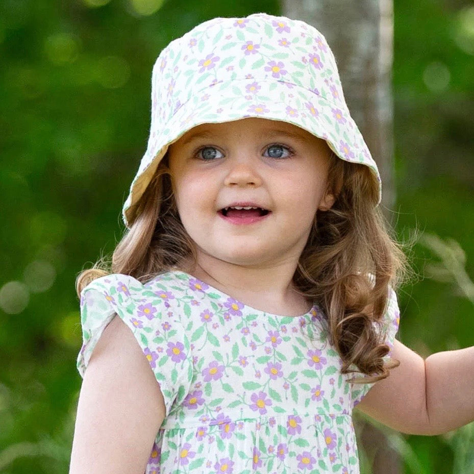 A young child wearing a white sun hat with a green and purple floral print, paired with a matching floral dress, set against a soft, blurred green outdoor background.

Childrens hat
Sun hat 
Kids clothing 
Floral print
Reversible 