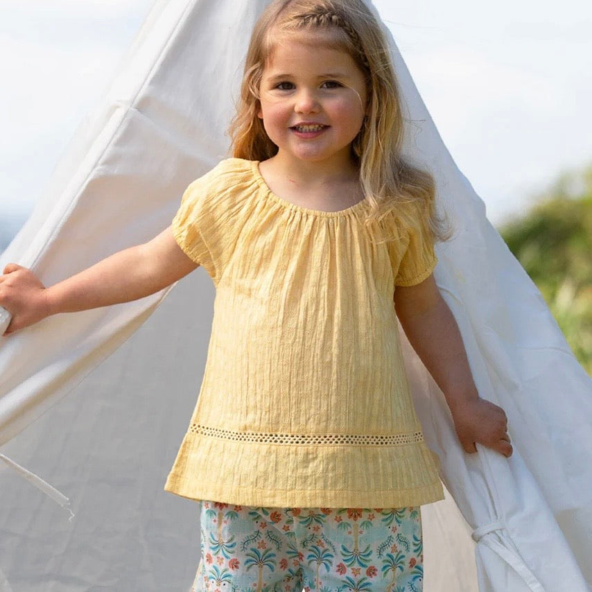 Young girl in a yellow top and floral pants standing inside a white tent.

Children's clothing 
Yellow
Blouse
Organic cotton 
Summer clothing 
Summer