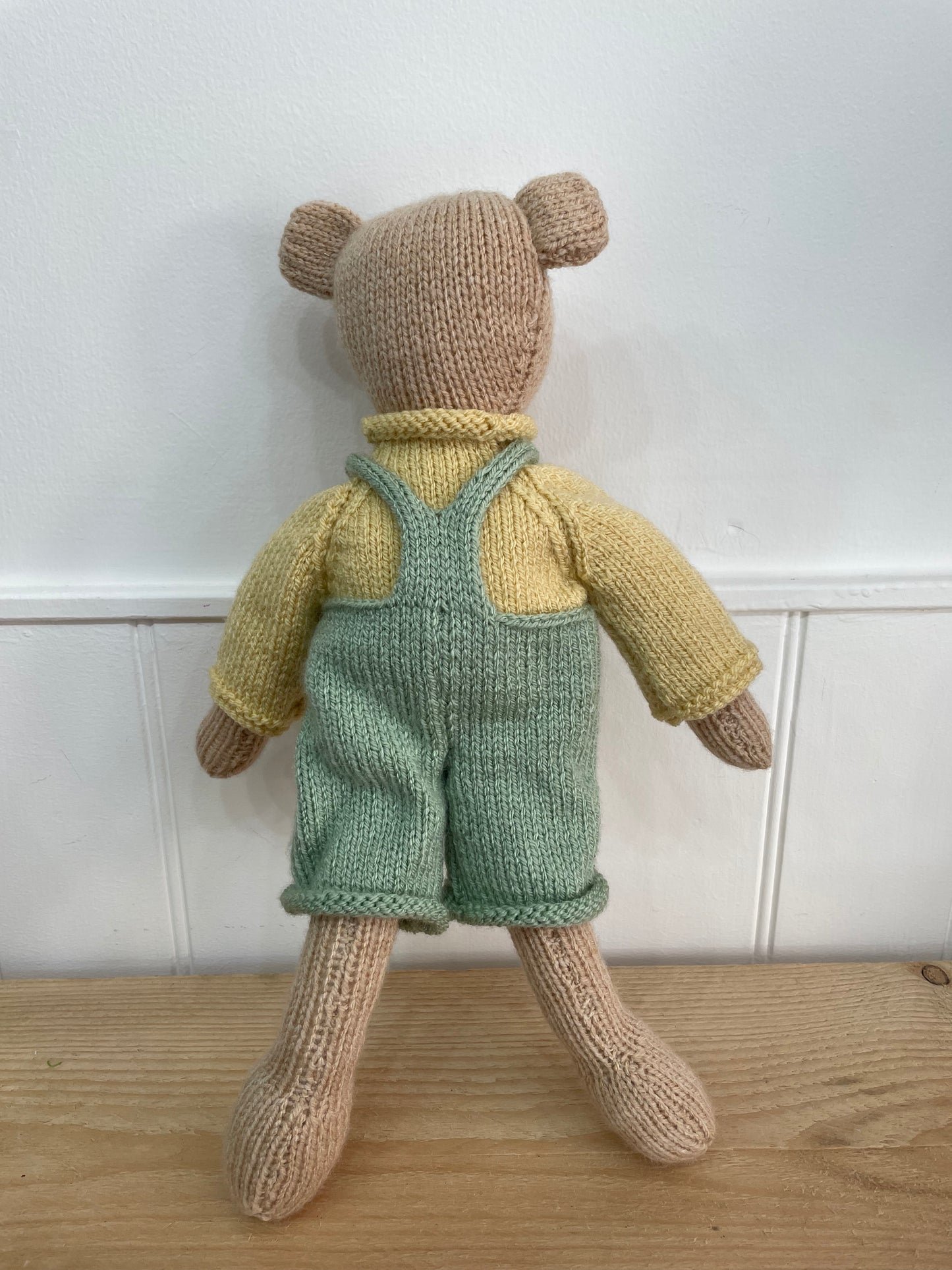 Knitted teddy bear wearing a yellow shirt and green overalls on a wooden surface with a white wall background.
A lovely present for a new baby