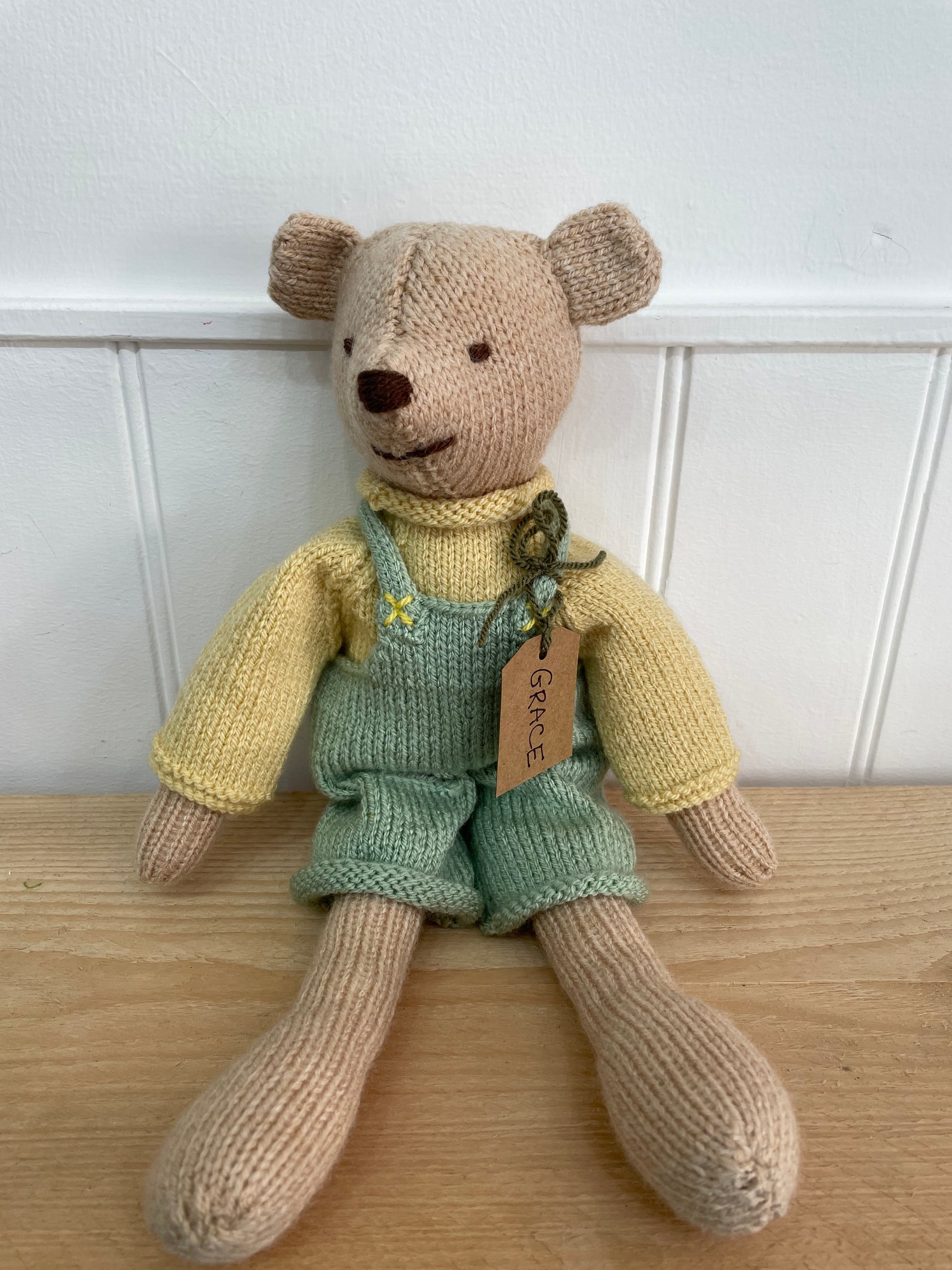 Knitted teddy bear wearing a yellow sweater and green overalls on a wooden surface with a white paneled wall background.
A lovely new baby present 