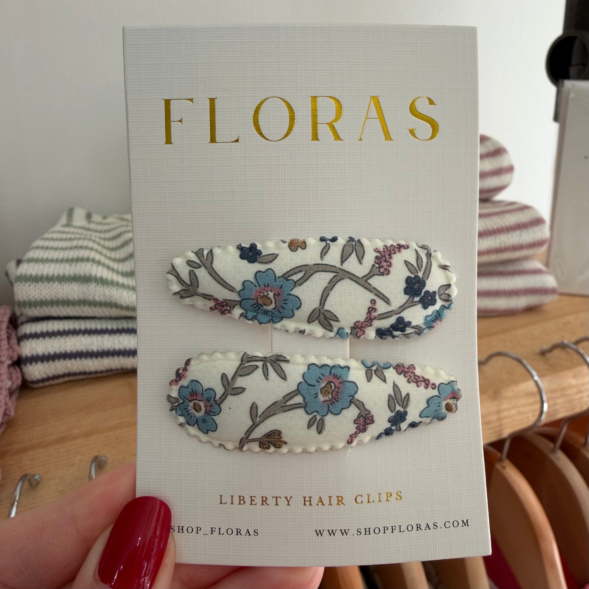 A hand with red nail polish holds a white display card featuring two floral embroidered hair clips. The card is branded 'FLORAS' in gold lettering and reads 'LIBERTY HAIR CLIPS'.

Hair Clips
Liberty Print
Gift inspiration 
Floral
Girls Hair Accessories