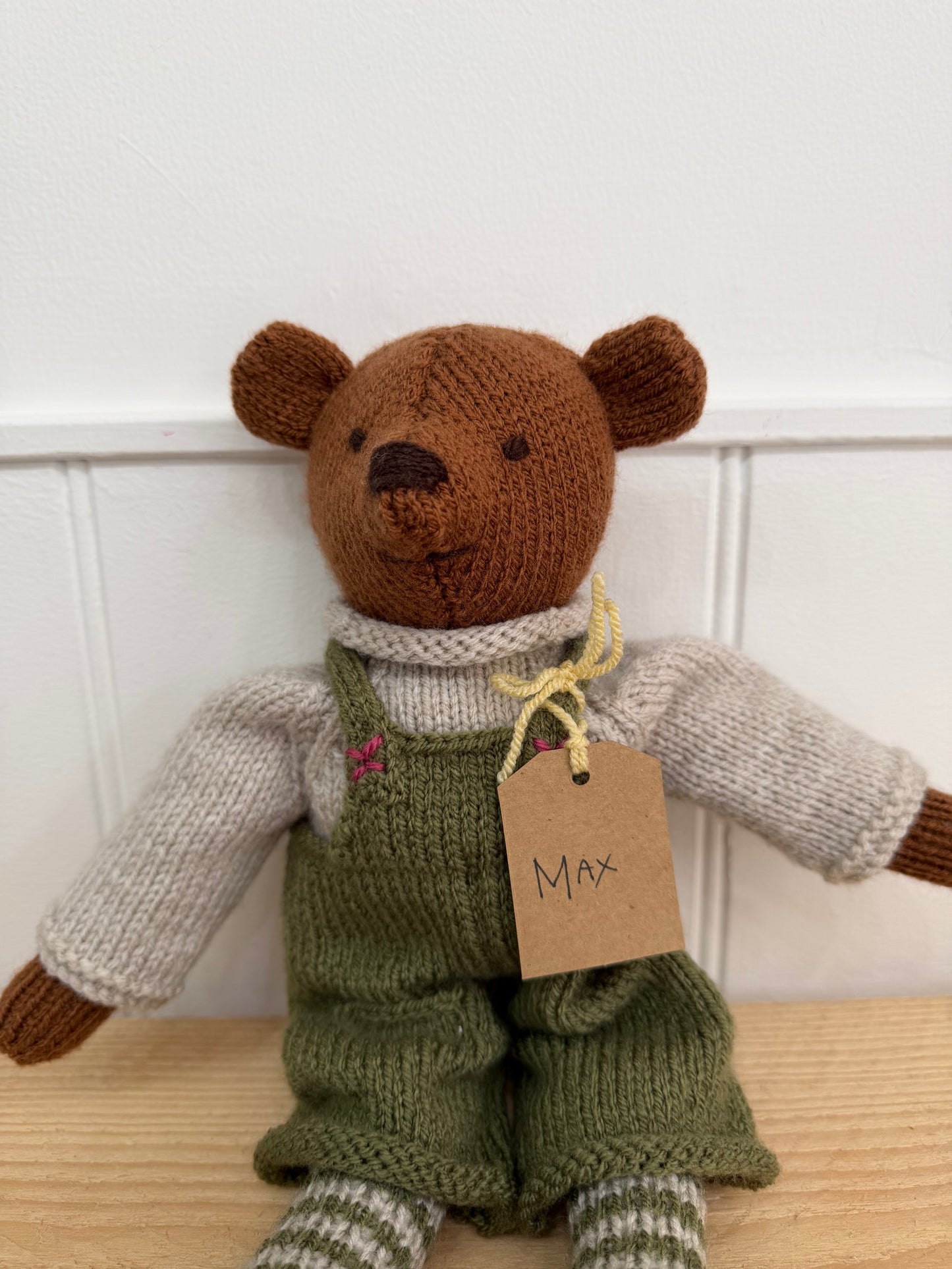 A handmade knitted teddy bear named Max, wearing an olive green knitted overall, a cream-colored knit sweater, and striped socks, sitting on a light wooden surface against a white panelled wall.

Hand knitted bear 
Perfect for new baby presents
Locally made
Hand made
Unique presents 