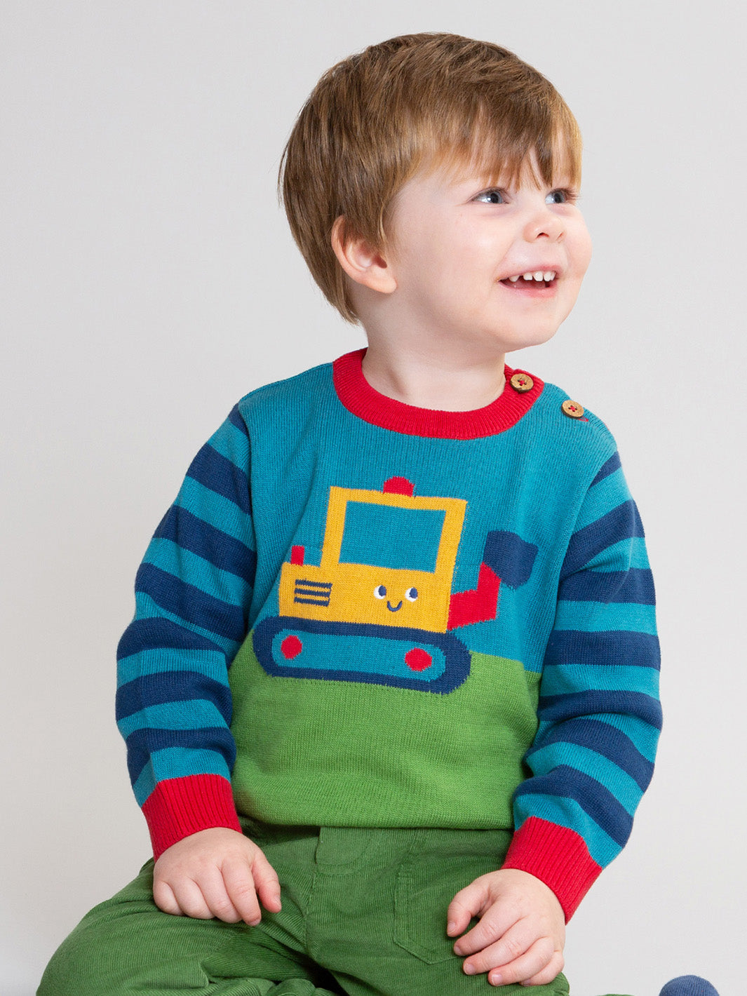 A smiling young boy wearing a colourful knit sweater with a yellow construction vehicle graphic, blue and teal horizontal stripes on the sleeves, and a green body, paired with green pants, photographed against a plain light grey background.

Children's clothing
Jumper
Digger
Boys clothing
Kite clothing 
Organic cotton 
Cosy cords 