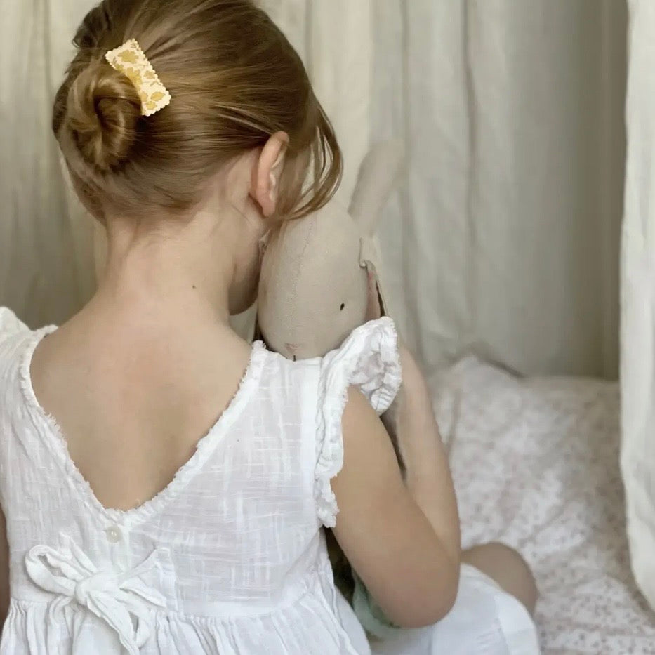 A young girl with blonde hair in a bun, wearing a white ruffled dress and holding a plush bunny, shown from behind in a soft, neutral-toned bedroom setting.

Hair Clips
Liberty Print
Gift inspiration 
Floral
Girls Hair Accessories