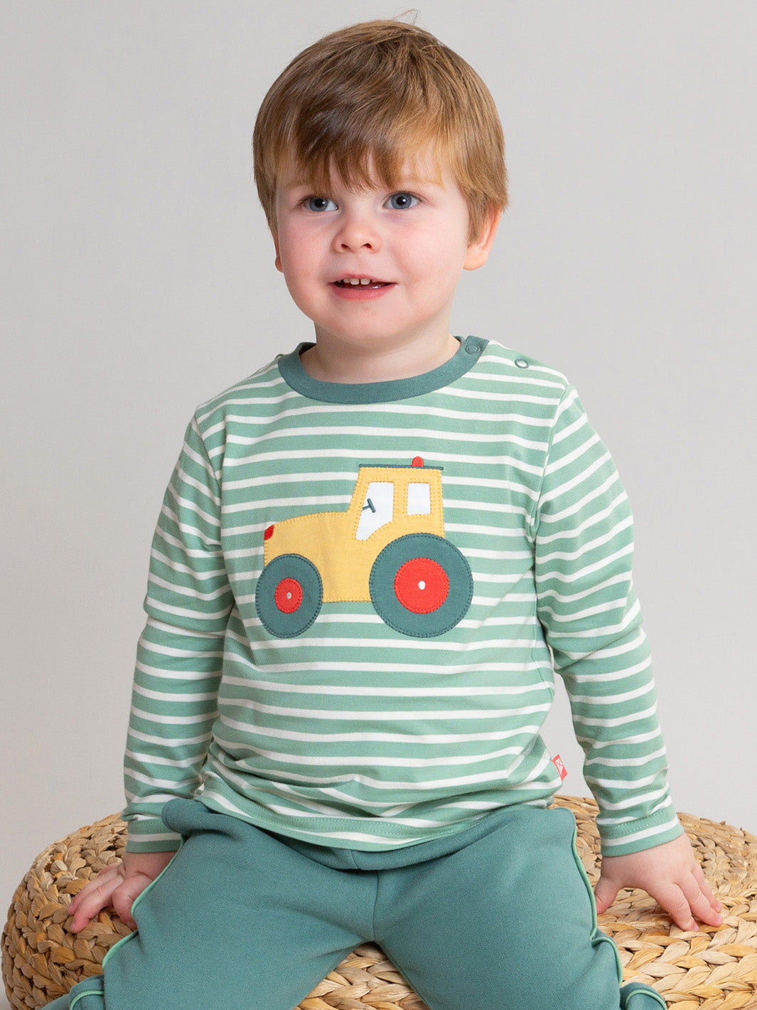 A toddler boy wearing a green and white striped long-sleeve shirt with a yellow tractor graphic on the front, paired with matching green pants, sitting on a woven basket against a light gray background.

Children's clothing 
Organic cotton 
T-Shirt
Tractors 
Farm 
Autumn clothing
