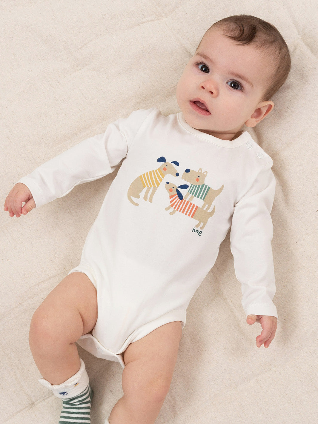 A baby lying on a cream-coloured textured blanket, wearing a white long-sleeve bodysuit with a colourful cartoon dog graphic on the chest and striped socks.

Baby clothing 
Kite clothing
Organic cotton 
New baby gift
Gender neutral 