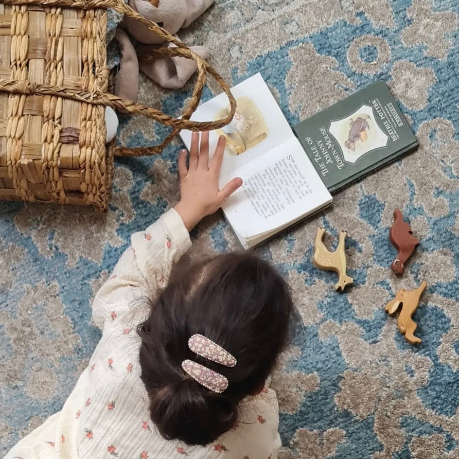 A child seen from above wearing a cream floral long-sleeve shirt and two pink rhinestone hair clips, sitting on a blue patterned rug next to a woven basket, an open book, and wooden animal toys.

Hair Clips
Liberty Print
Gift inspiration 
Floral
Girls Hair Accessories