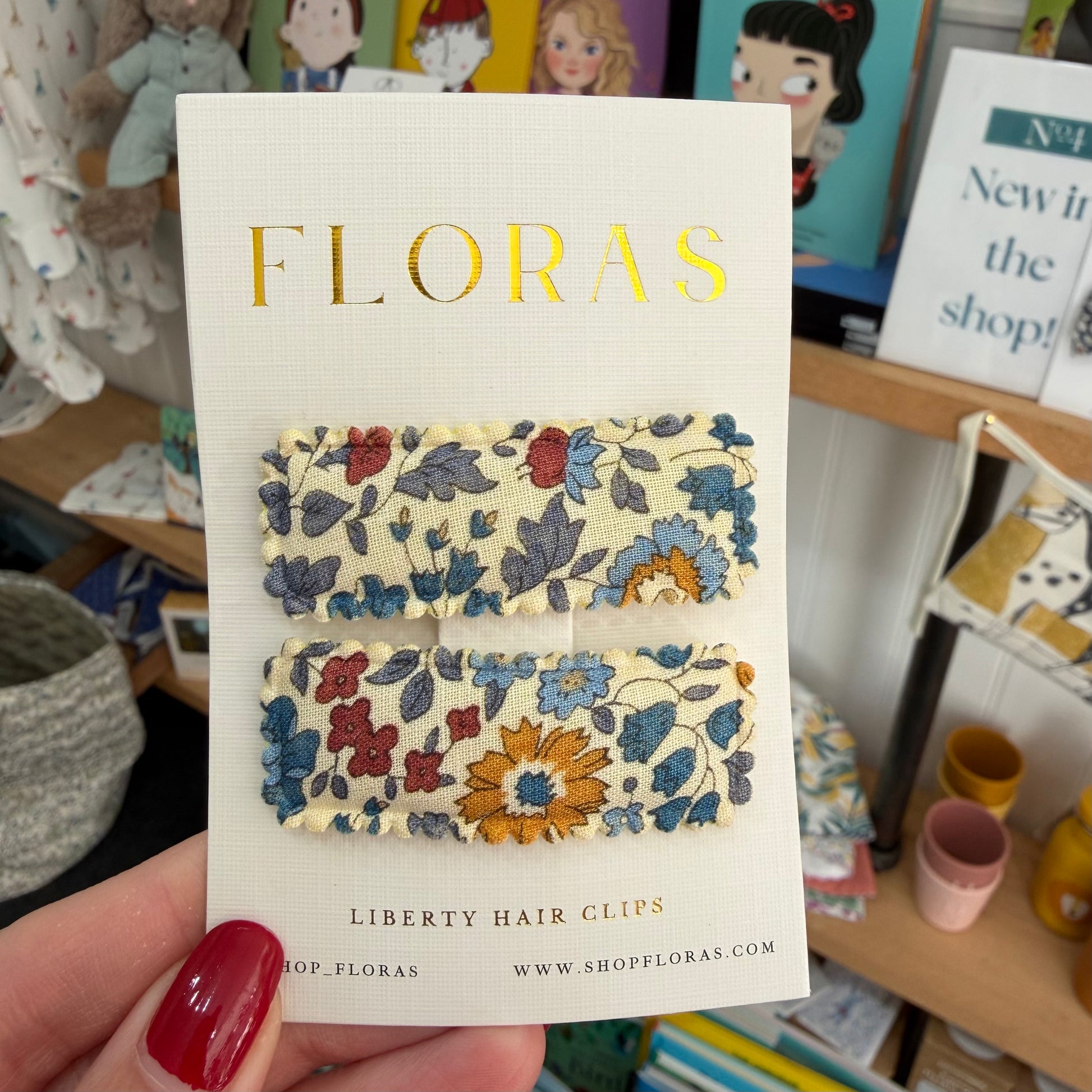 Two Liberty hair clips with a floral pattern on a cream background, presented on a white card branded 'FLORAS' held by a hand with red nail polish in a shop setting.

Hair Clips
Liberty Print
Gift inspiration 
Floral
Girls Hair Accessories