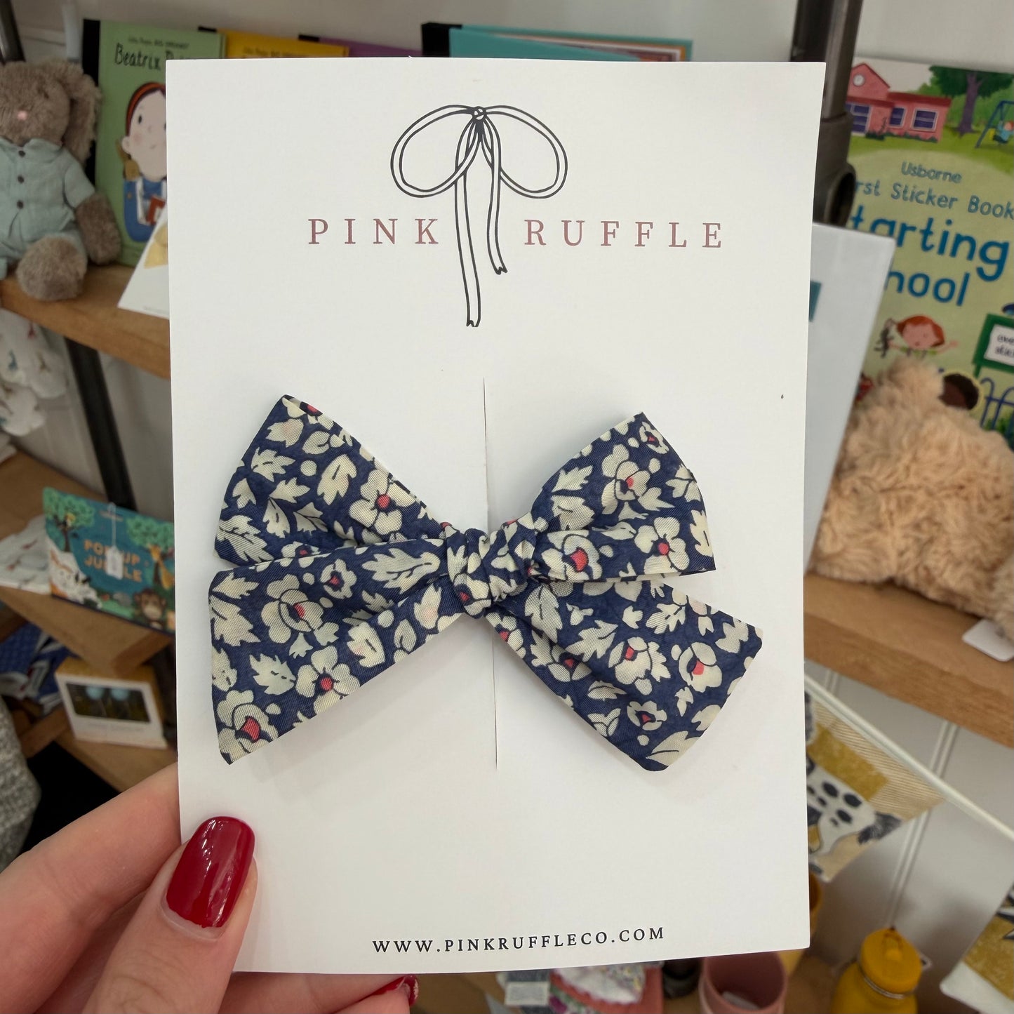A navy blue bow with a cream and red floral print, displayed on a white 'Pink Ruffle' brand card held by a hand with red nail polish.

Hair Clips
Liberty Print
Gift inspiration 
Floral
Girls Hair Accessories