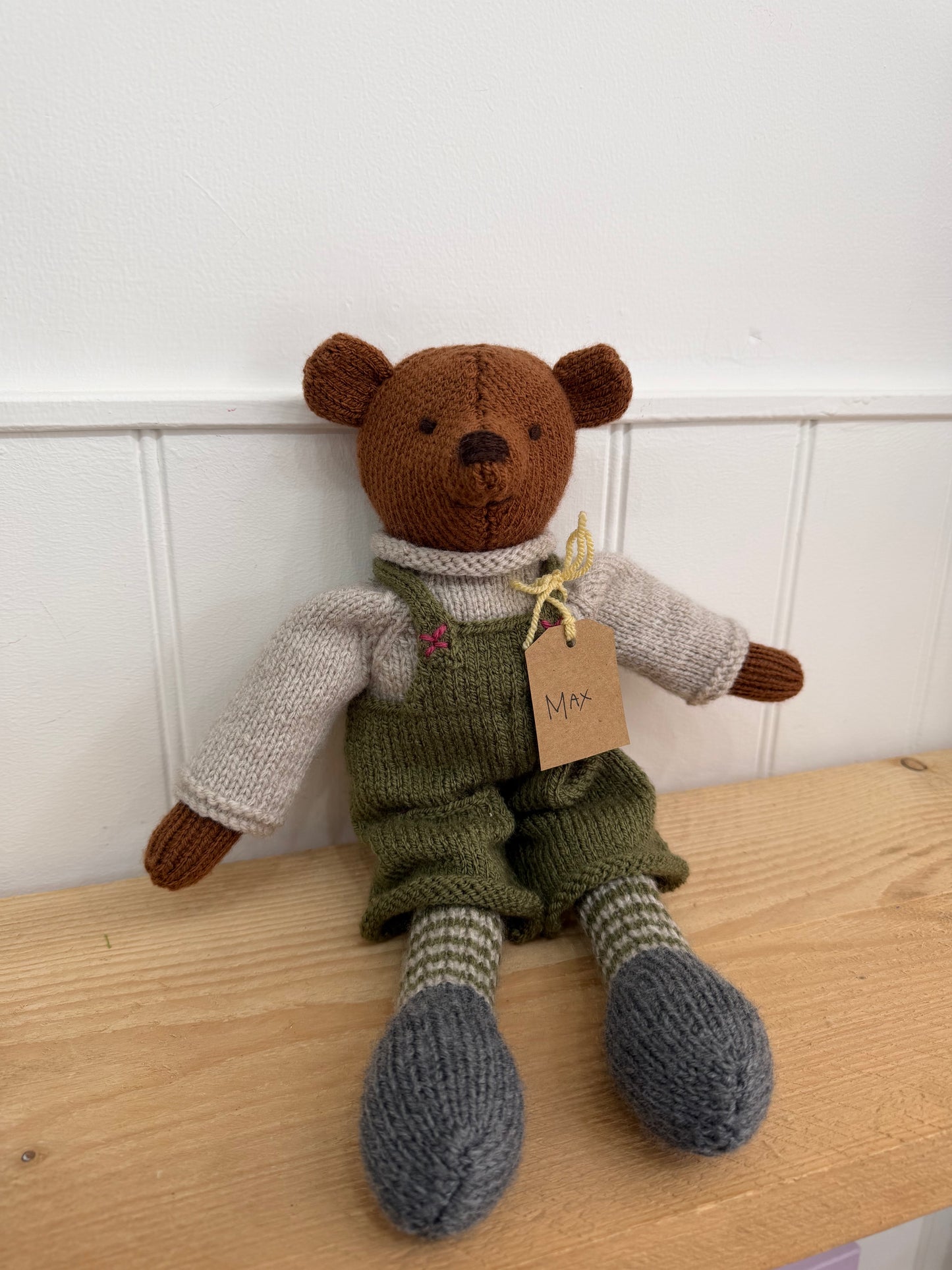 A handmade knitted teddy bear sitting on a light wooden table against a white panelled wall. The bear is brown with a cream knit sweater, olive green overalls, striped green and cream socks, and grey knitted shoes.

Hand knitted bear 
Perfect for new baby presents
Locally made
Hand made
Unique presents 