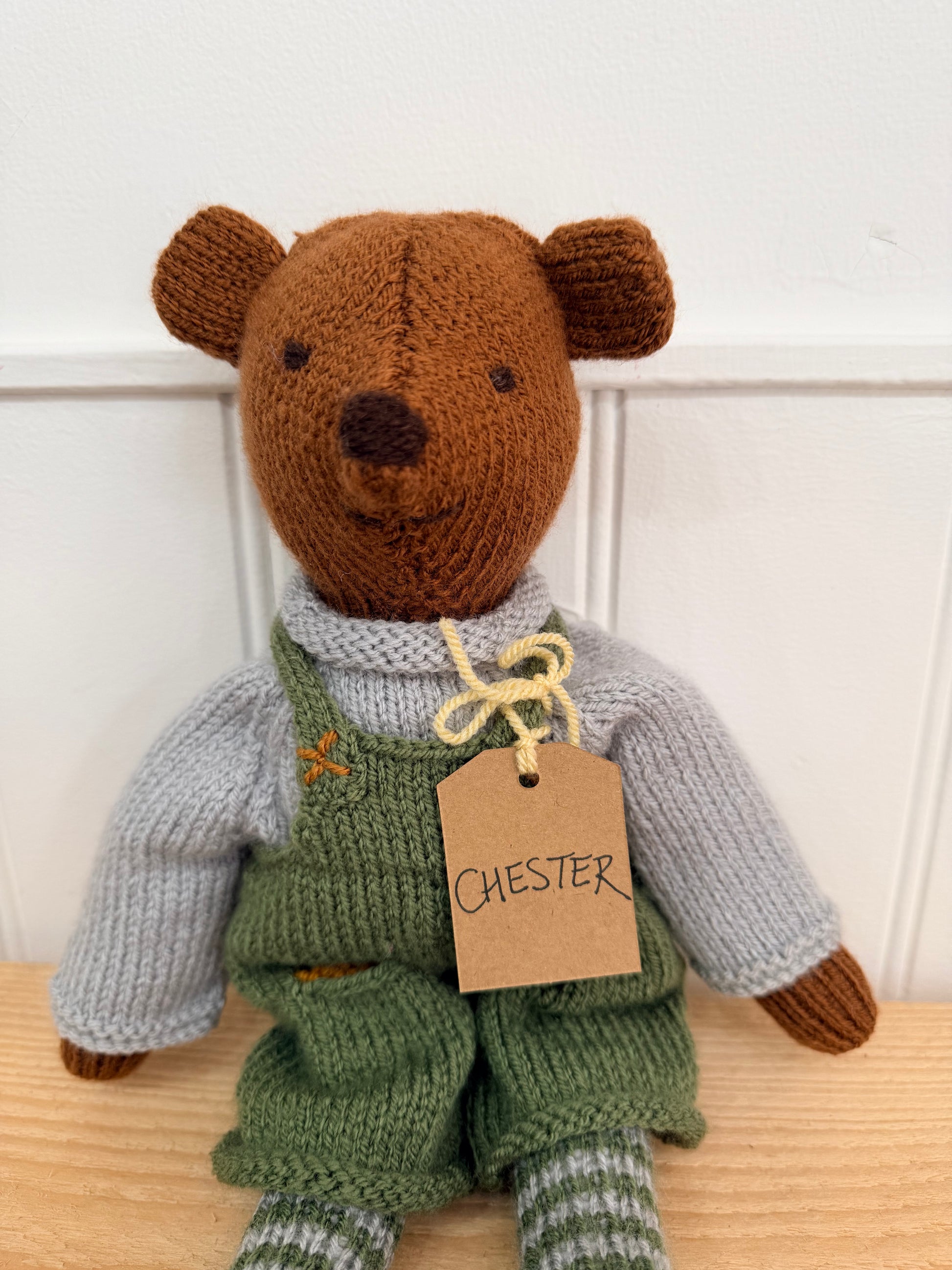 A handmade knitted teddy bear named Chester, wearing green overalls, a light grey sweater, and striped socks, sitting on a wooden surface against a white panelled wall.

Hand knitted bear 
Perfect for new baby presents
Locally made
Hand made
Unique presents 