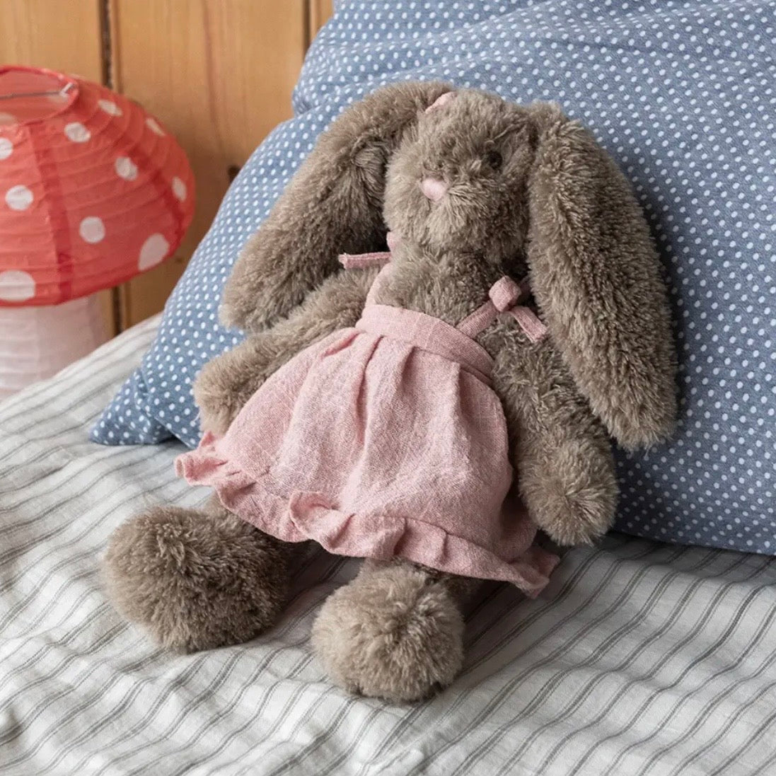 A plush gray bunny doll wearing a pink dress, posed on a bed with a blue polka-dot pillow and striped bedding in the background.

cuddly toy rabbit, soft toy for girls, bunny stuffed animal, nursery plush toy, Easter bunny plush, comforter toy, huggable toy for toddlers, baby shower gift, keepsake soft toy, rabbit plushie, dressed bunny toy
