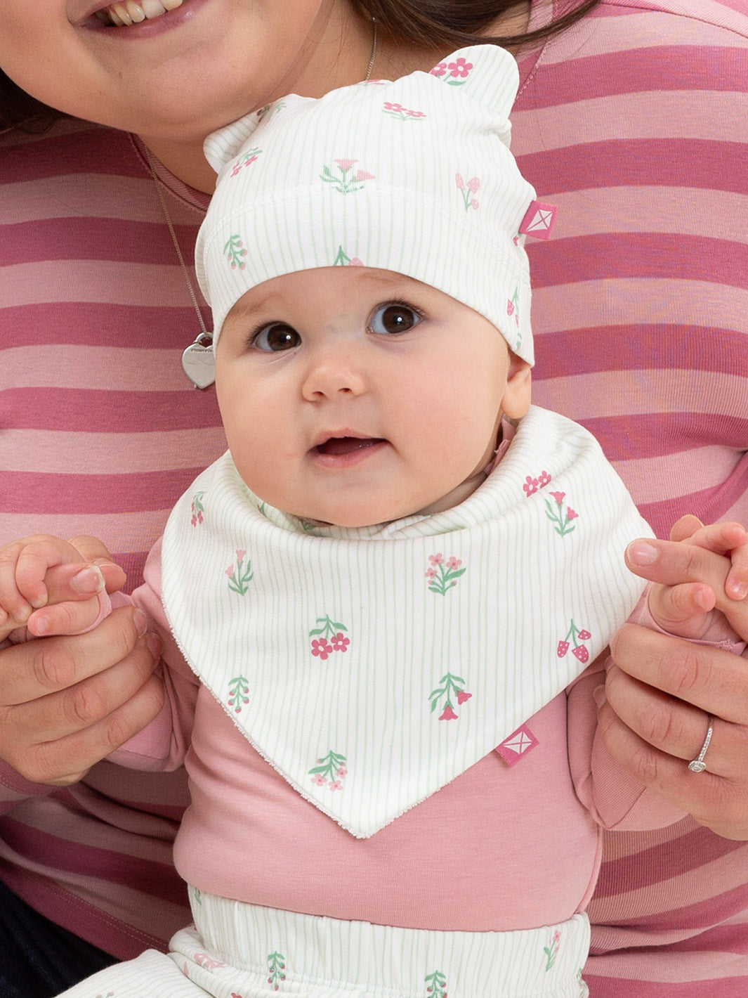 A baby wearing a white hat and matching bib with a small pink floral print, held by an adult wearing a pink and white striped shirt.

New baby gifts
Bib
Floral
Organic cotton
Kite clothing