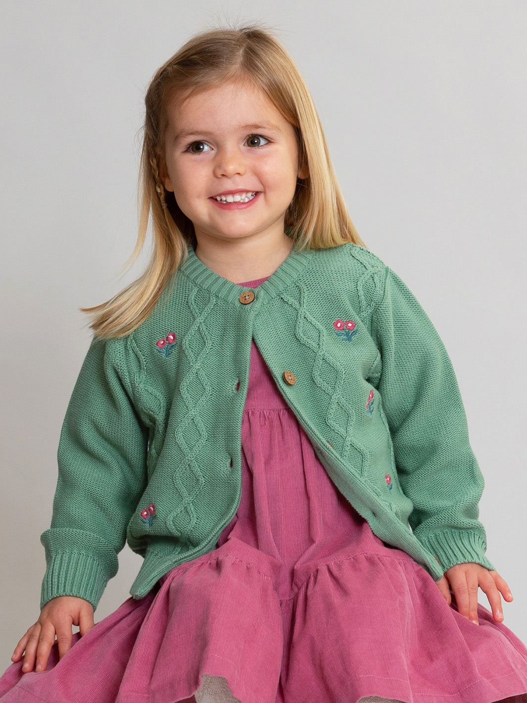 A young girl with blonde hair smiling, wearing a sage green cable-knit cardigan with embroidered flowers and a pink tiered dress, set against a plain light gray studio background.

Girls clothing
Children's clothing
Cardi
Floral
Organic cotton 
Kite clothing