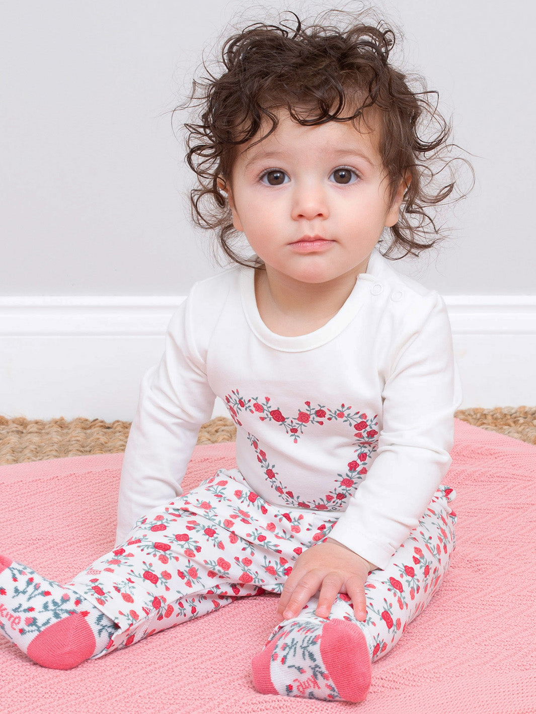 Child wearing a floral outfit sitting on a pink blanket.