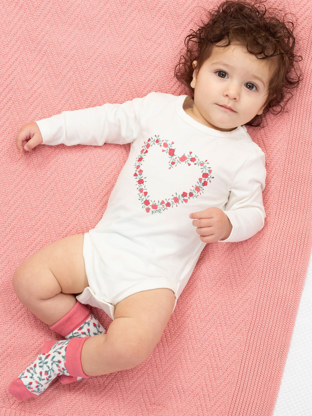 Baby wearing a white onesie with a floral design on a pink blanket with rose socks