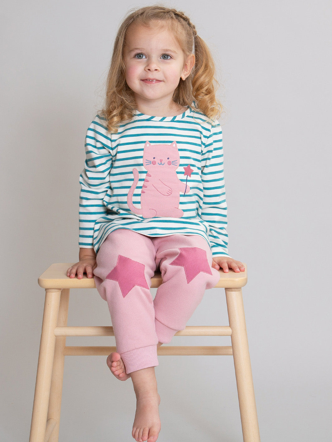 A young girl with blonde pigtails sits on a wooden stool wearing a long-sleeve pajama set with teal and white stripes, a pink cat graphic, and pink joggers with star patches.

Children's clothing
Organic cotton 
Kids clothing
Kite clothing
Joggers
Star