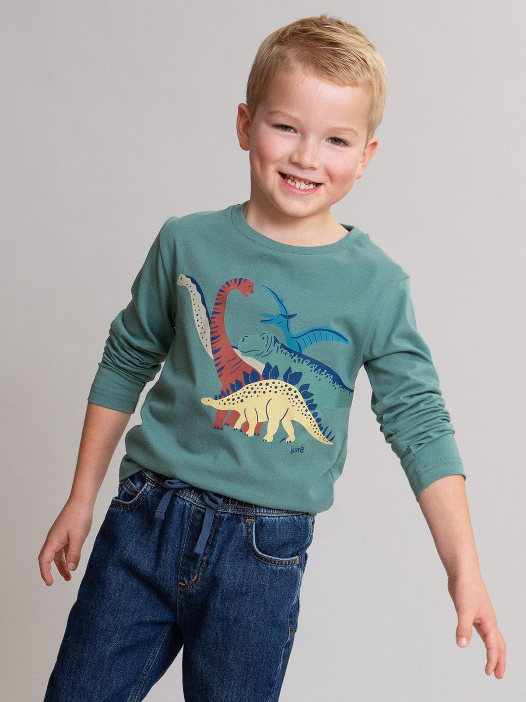 A smiling young boy wearing a sage green long-sleeve crewneck shirt with a colorful dinosaur graphic on the front, paired with blue jeans, against a solid light gray background.

Children's clothing
Dino
Organic cotton 
T-shirt
Boys clothing 
Kite clothing 