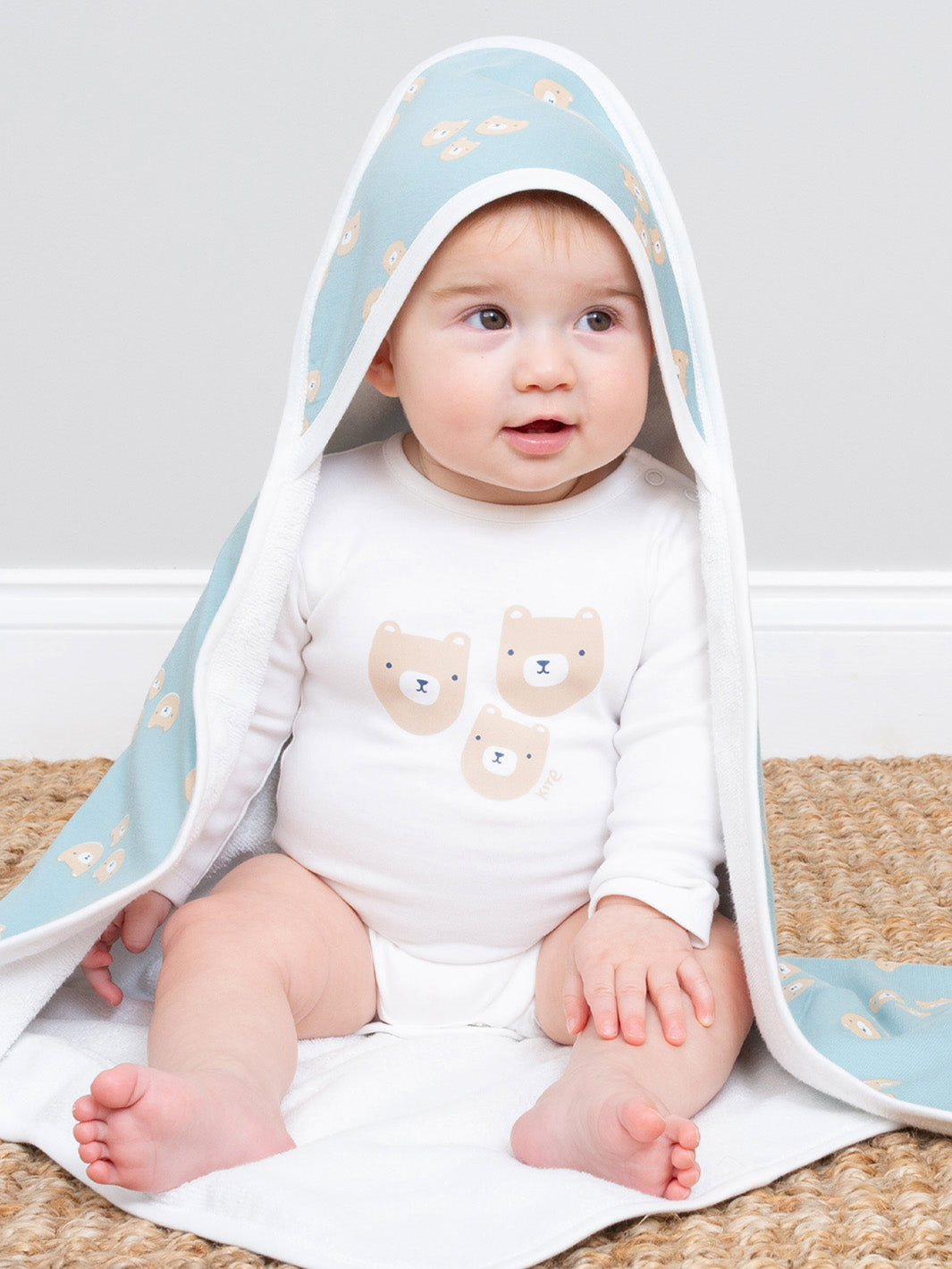 Baby sitting on a rug with a blue hooded towel draped over them, featuring bear designs.