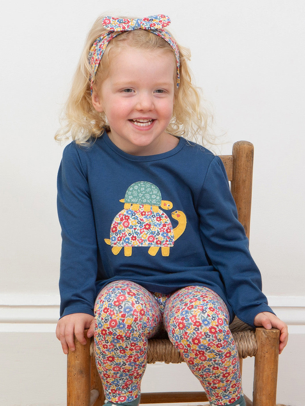 Child wearing a blue tortoise tunic and floral leggings, sitting on a wooden chair.