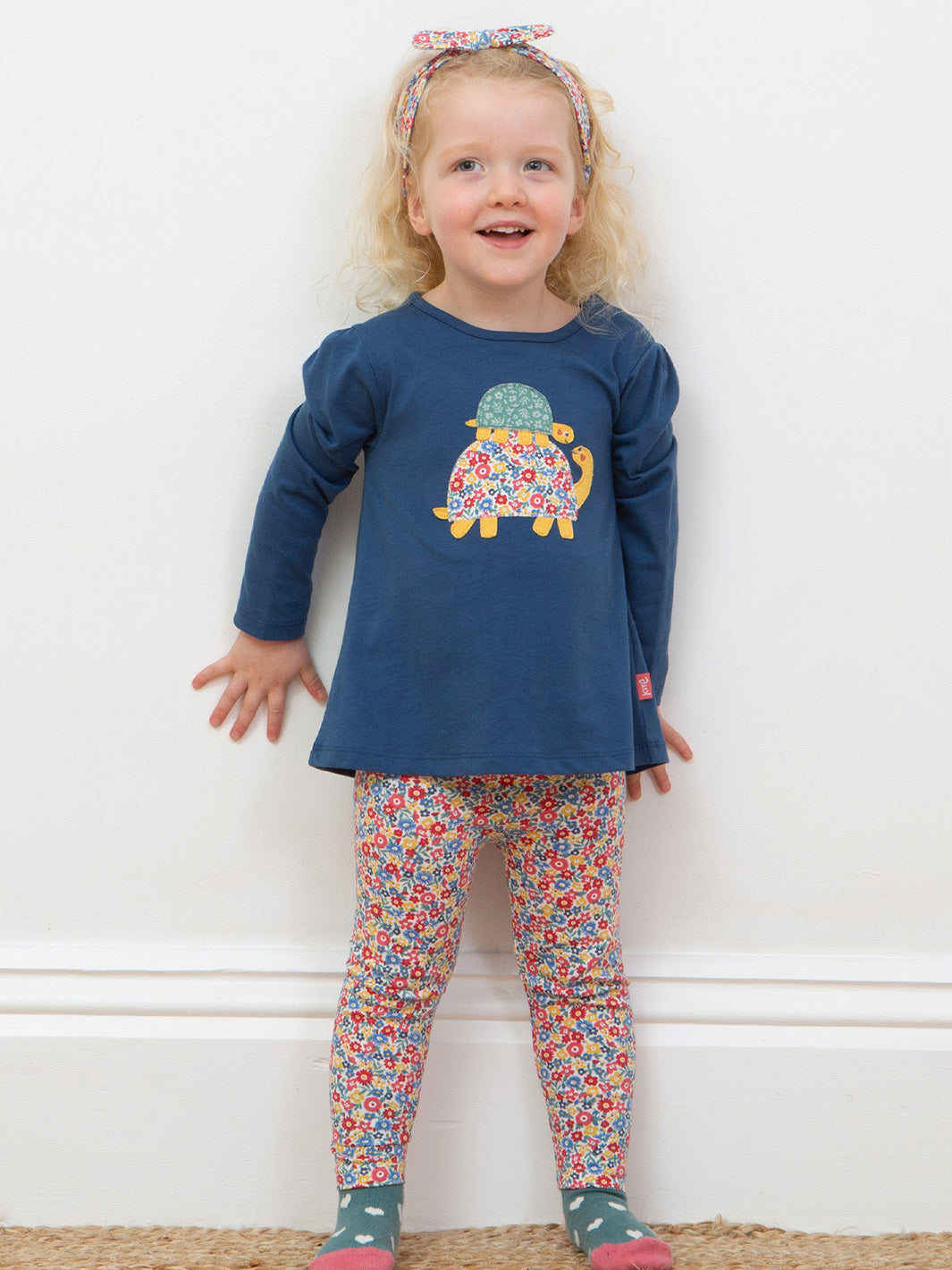 Child wearing a blue top with a turtle design and colorful floral leggings against a white wall.