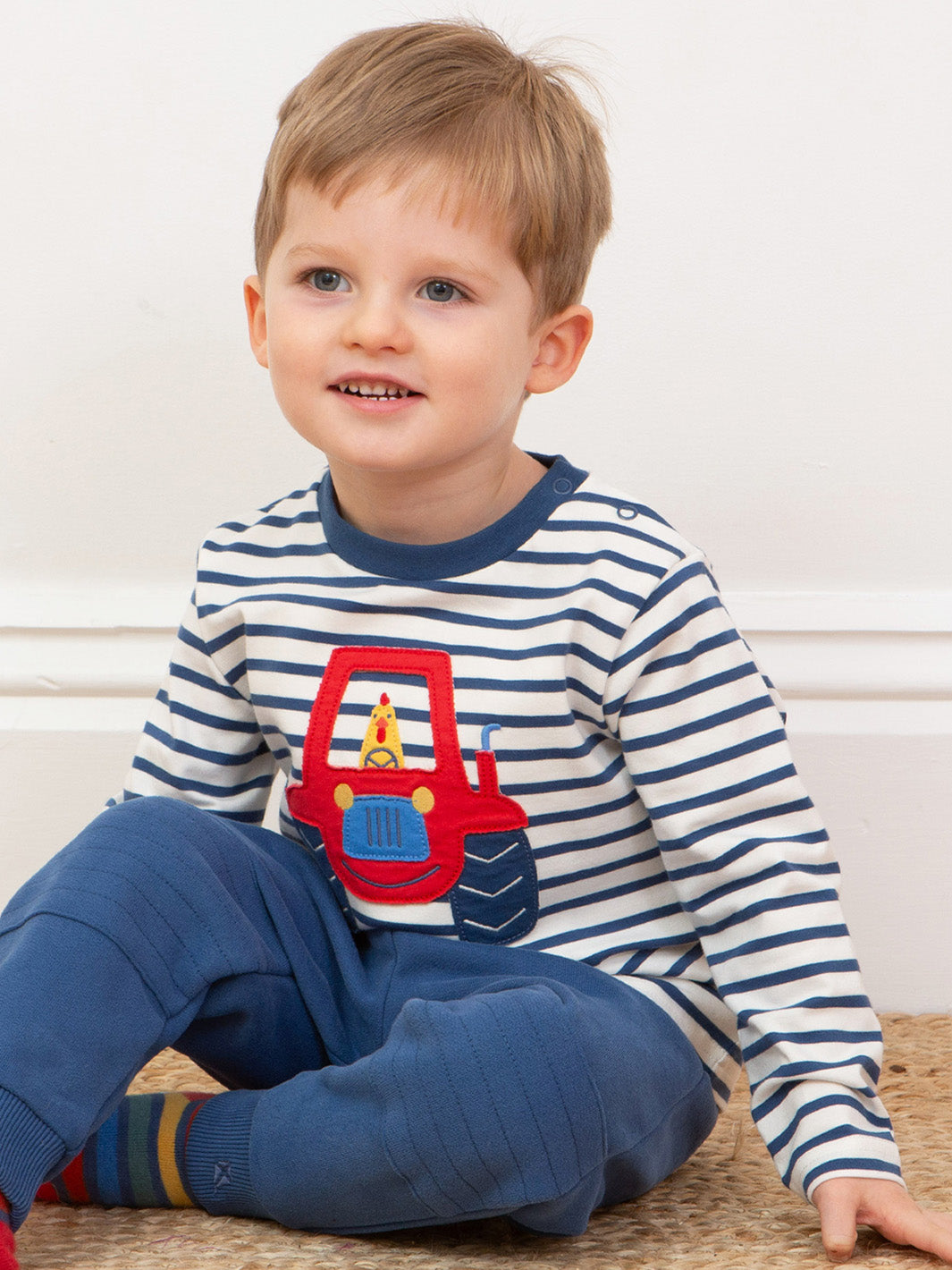Child wearing a striped shirt with a red tractor design, sitting on a wooden floor.