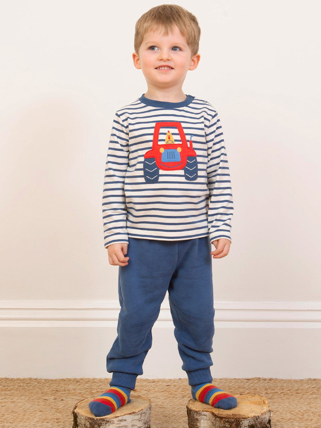 Child wearing a striped shirt with a red tractor design and blue trousers on a wooden platform.