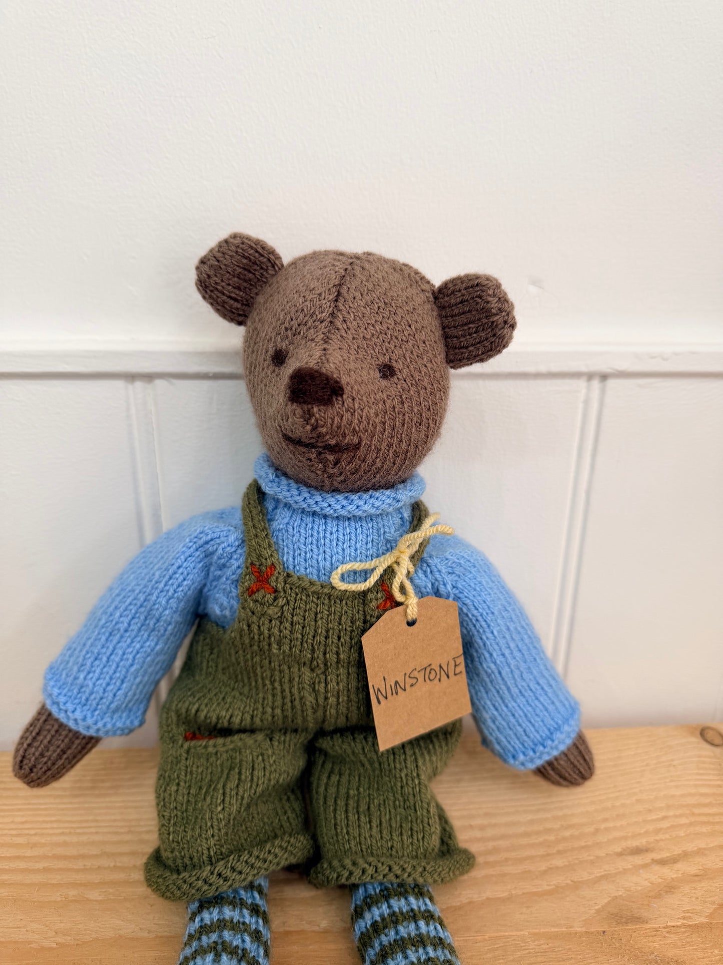 A handmade knitted teddy bear doll wearing a light blue turtleneck sweater and olive green overalls, sitting on a light wood surface against a white paneled wall. A small paper tag is attached to the neck and reads 'WINSTONE'.

Hand knitted bear 
Perfect for new baby presents
Locally made
Hand made
Unique presents 