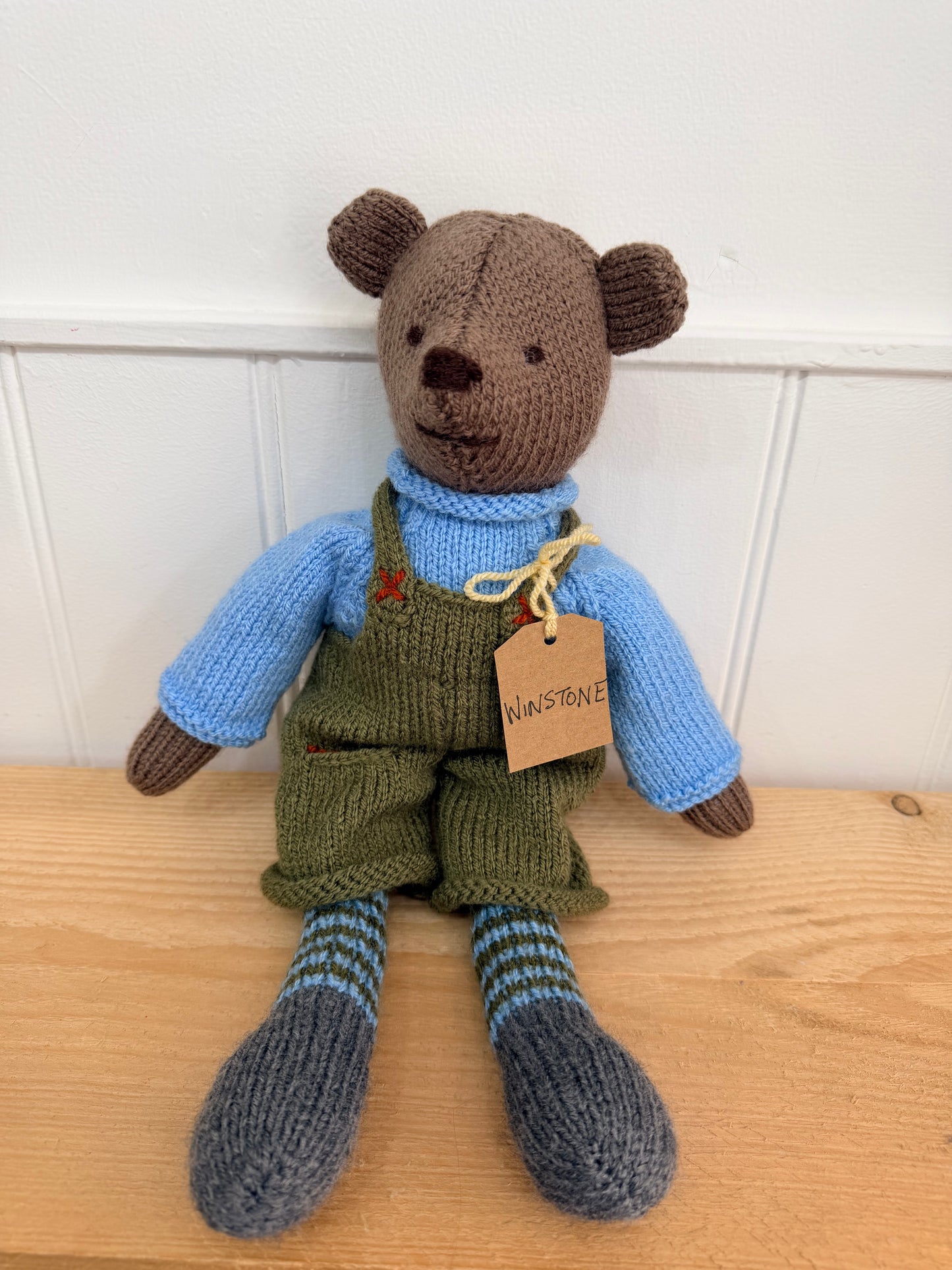 A handmade knitted teddy bear sitting on a light wooden surface against a white panelled wall. The bear is brown with a blue knit shirt, olive green overalls, and blue and gray striped socks.

Hand knitted bear 
Perfect for new baby presents
Locally made
Hand made
Unique presents 