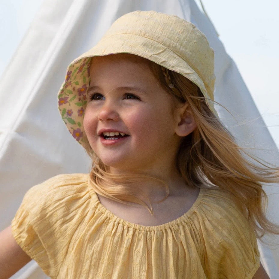 A young girl smiling outdoors while wearing a pale yellow sun hat with a floral patterned brim and a matching pale yellow top.

Childrens hat
Sun hat 
Kids clothing 
Floral print
Reversible 